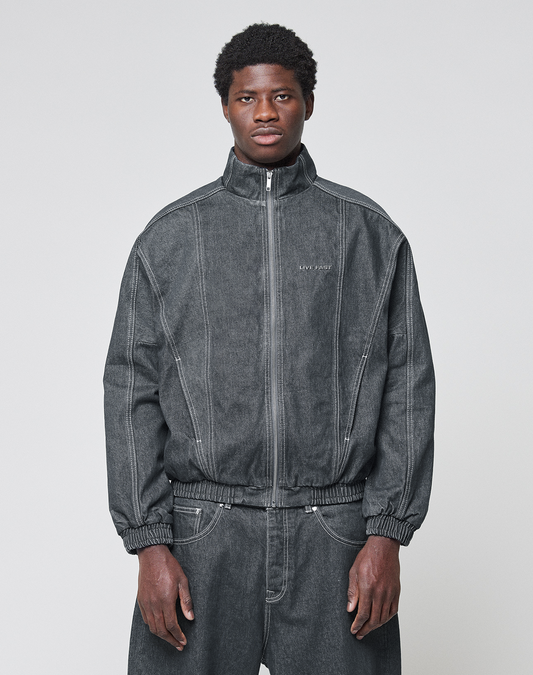 A man faces forward in a dark gray LFDY Denim Trackjacket by LIVE FAST DIE YOUNG, with visible seams, subtle chest text, and paired baggy pants. The 100% cotton jacket features a stand-up collar and is set against a plain light background. | model
