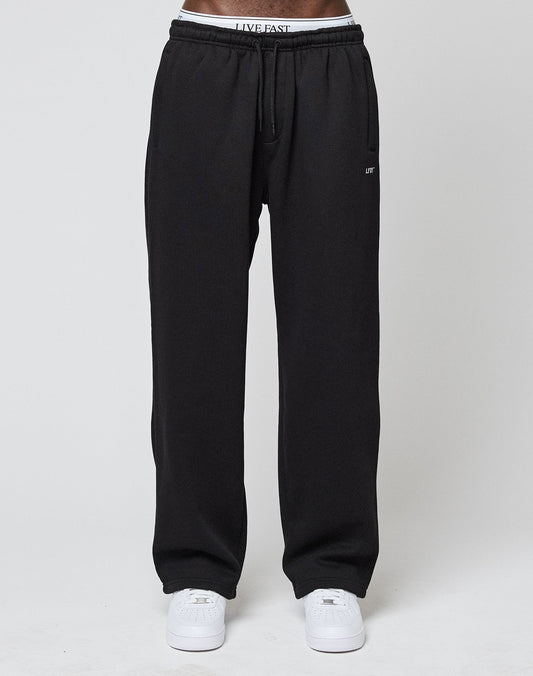 A person wears LIVE FAST DIE YOUNG Basic Wide Jogger sweatpants with an elastic waistband and logo print, partially revealing white underwear with LIVE FAST lettering, plus white sneakers, standing against a plain light background.