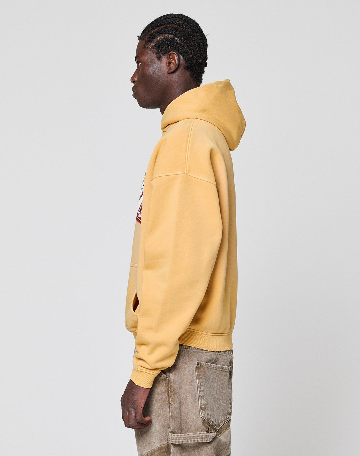 A person with braided hair stands in profile wearing the LIVE FAST DIE YOUNG LFDY Corp. Hooded in yellow with a spray effect front print and light brown pants against a plain gray background.