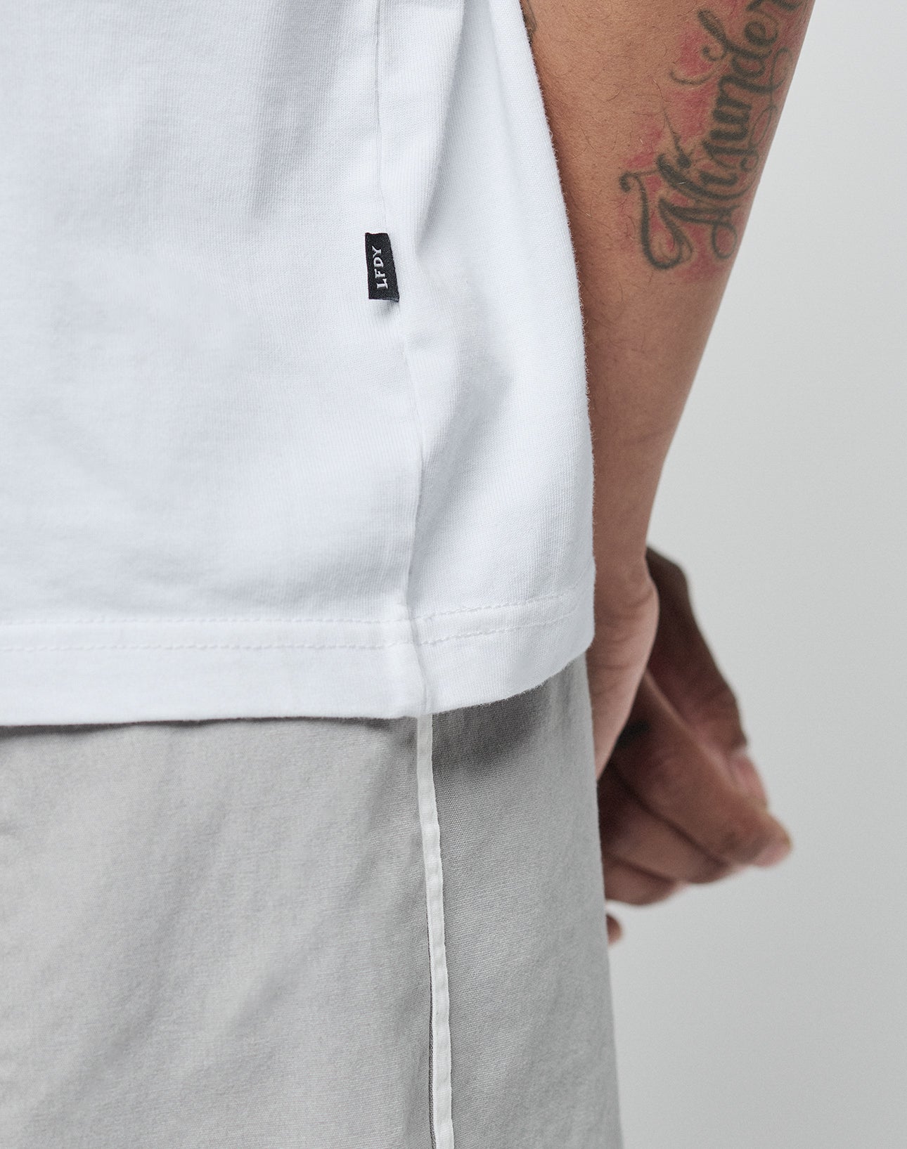 A close-up of a person in the LIVE FAST DIE YOUNG Basic +49 Tee in white, made from weicher Baumwolle, and light gray shorts. The shirt has a black tag; a forearm tattoo is visible as their hand rests at their side.