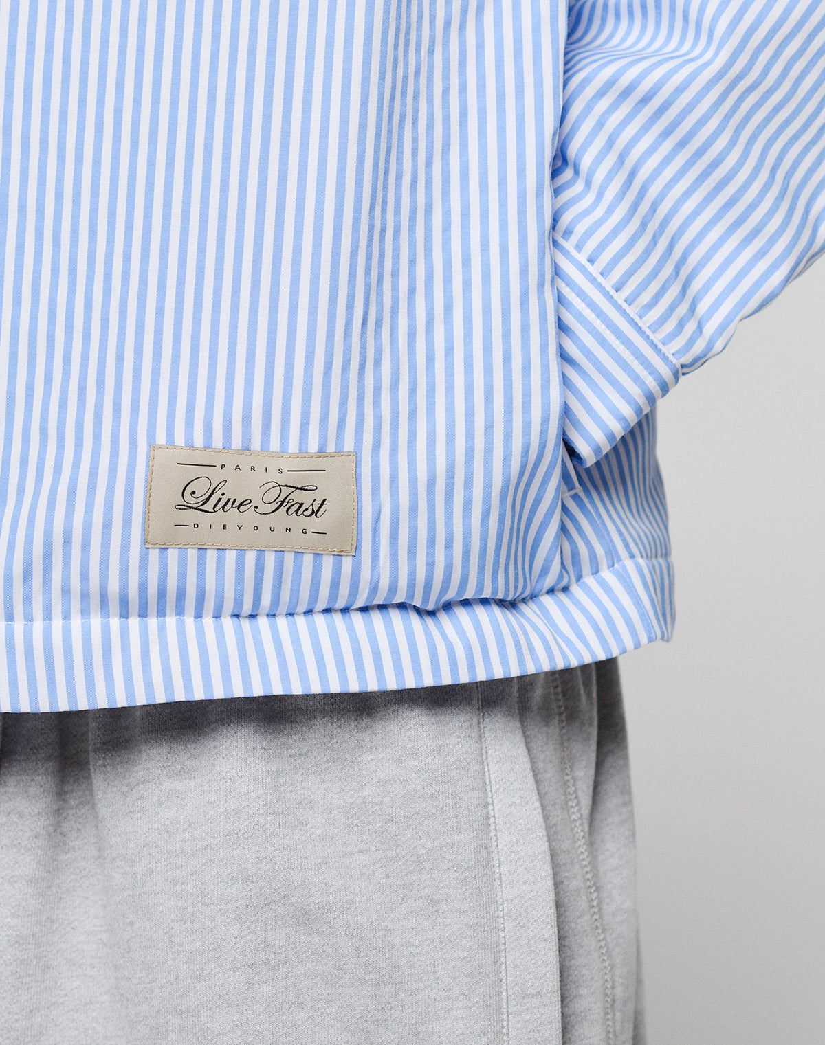 Close-up of a person wearing the LFDY Striped Padded Shirt by LIVE FAST DIE YOUNG, featuring blue and white vertical stripes, brusttaschen, and a beige "Live Fast" patch, paired with light gray sweatpants.