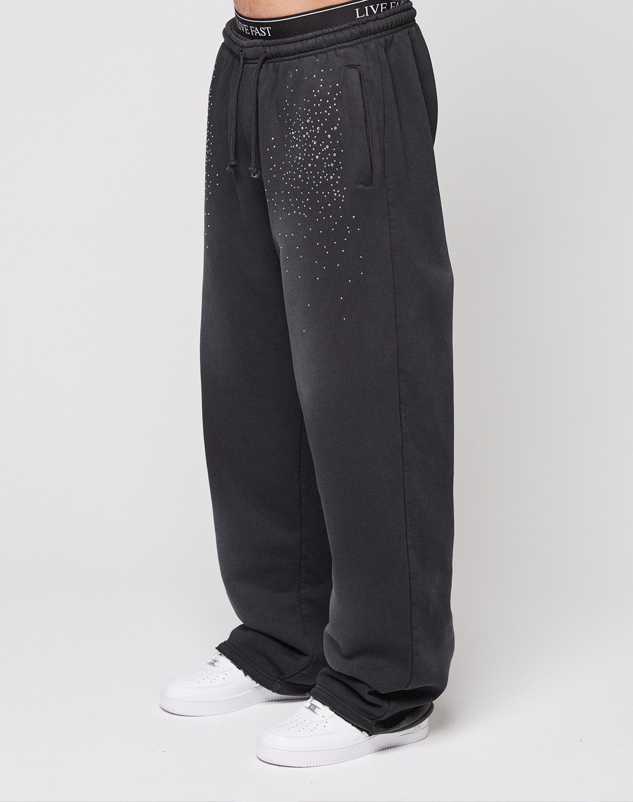 A person wears the LIVE FAST DIE YOUNG Live Fast Rhinestone Jogger, a wide fit black sweatpant with front rhinestone detail, drawstring waist, and bold LIVE FAST waistband text, styled with white sneakers against a plain white background.