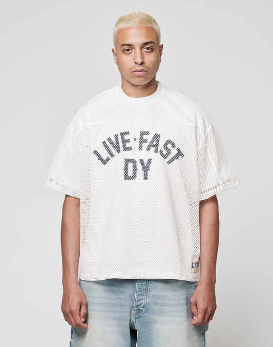 A person with short bleached blonde hair wears the LIVE FAST DIE YOUNG LFDY Boxy Jersey with a bold "LIVE FAST D/Y" front print and light blue denim shorts, standing against a plain light gray background. | model