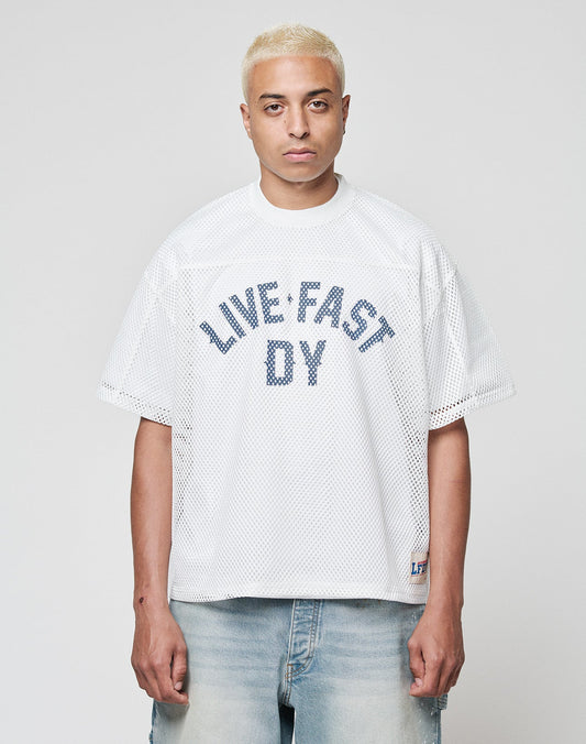 A person with short, bleached blond hair models the LIVE FAST DIE YOUNG LFDY Boxy Jersey—a loose white mesh shirt with a bold blue LIVE FAST DY front print—paired with light denim jeans, standing before a plain light gray backdrop.