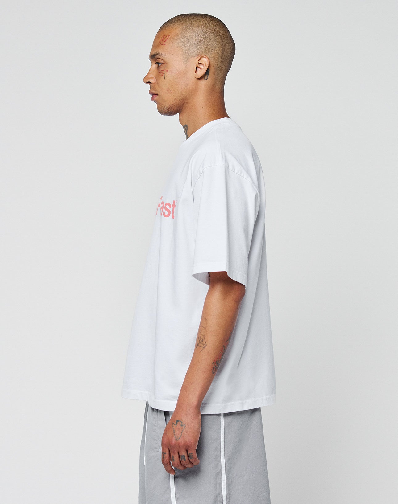 A person with a shaved head, visible tattoos, and wearing the LIVE FAST DIE YOUNG Basic +49 Tee in oversize fit and light gray shorts stands in profile against a plain white background.