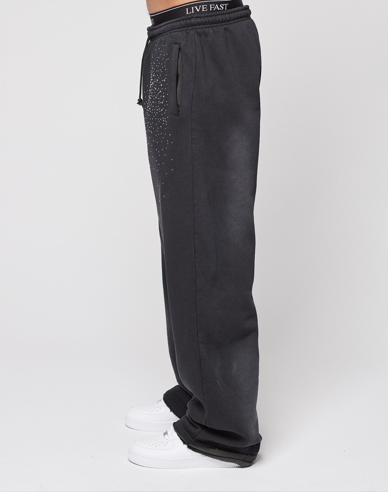 A person wears LIVE FAST DIE YOUNG Live Fast Rhinestone Jogger in wide fit black with a drawstring waist and white rhinestone Musters on the hip, plus a waistband that reads LIVE FAST. White sneakers complete the look against a plain white background.