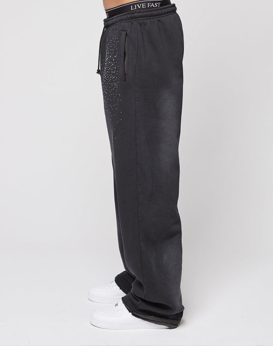 A person wears LIVE FAST DIE YOUNG Live Fast Rhinestone Jogger in wide fit black with a drawstring waist and white rhinestone Musters on the hip, plus a waistband that reads LIVE FAST. White sneakers complete the look against a plain white background.