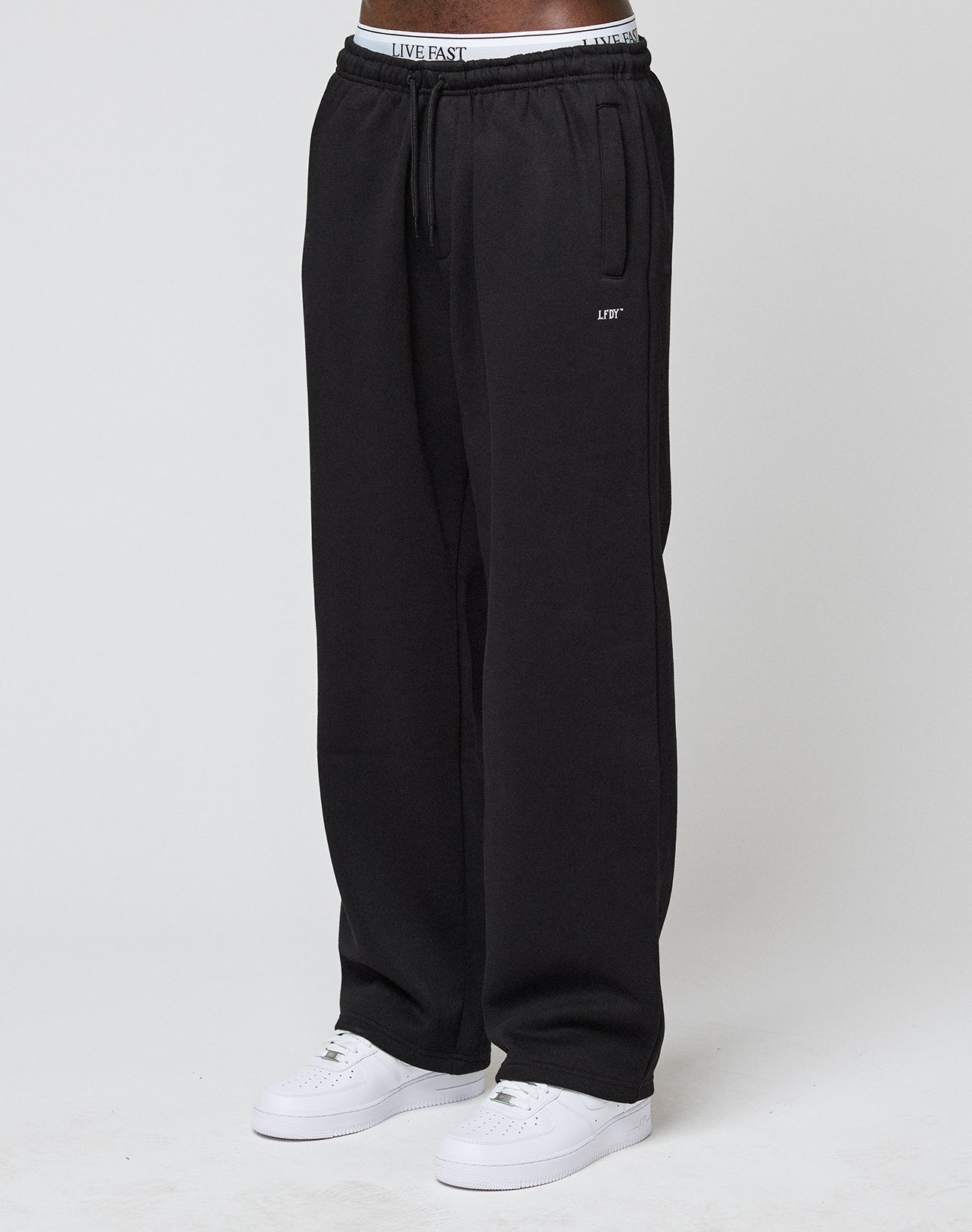 A person stands against a plain white background, wearing LIVE FAST DIE YOUNGs Basic Wide Jogger in black (loose fit), white sneakers, and a visible white waistband with the LIVE FAST logo. | model