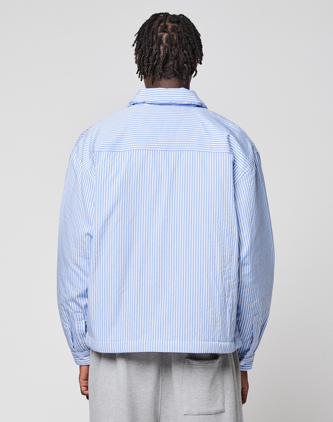 A person with short braids, seen from behind, wears the LFDY Striped Padded Shirt by LIVE FAST DIE YOUNG in light blue and white vertical stripes with brusttaschen and light gray pants against a plain, light background.