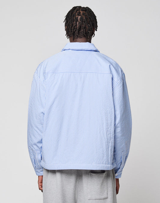 A person with short braids, seen from behind, wears the LFDY Striped Padded Shirt by LIVE FAST DIE YOUNG in light blue and white vertical stripes with brusttaschen and light gray pants against a plain, light background.