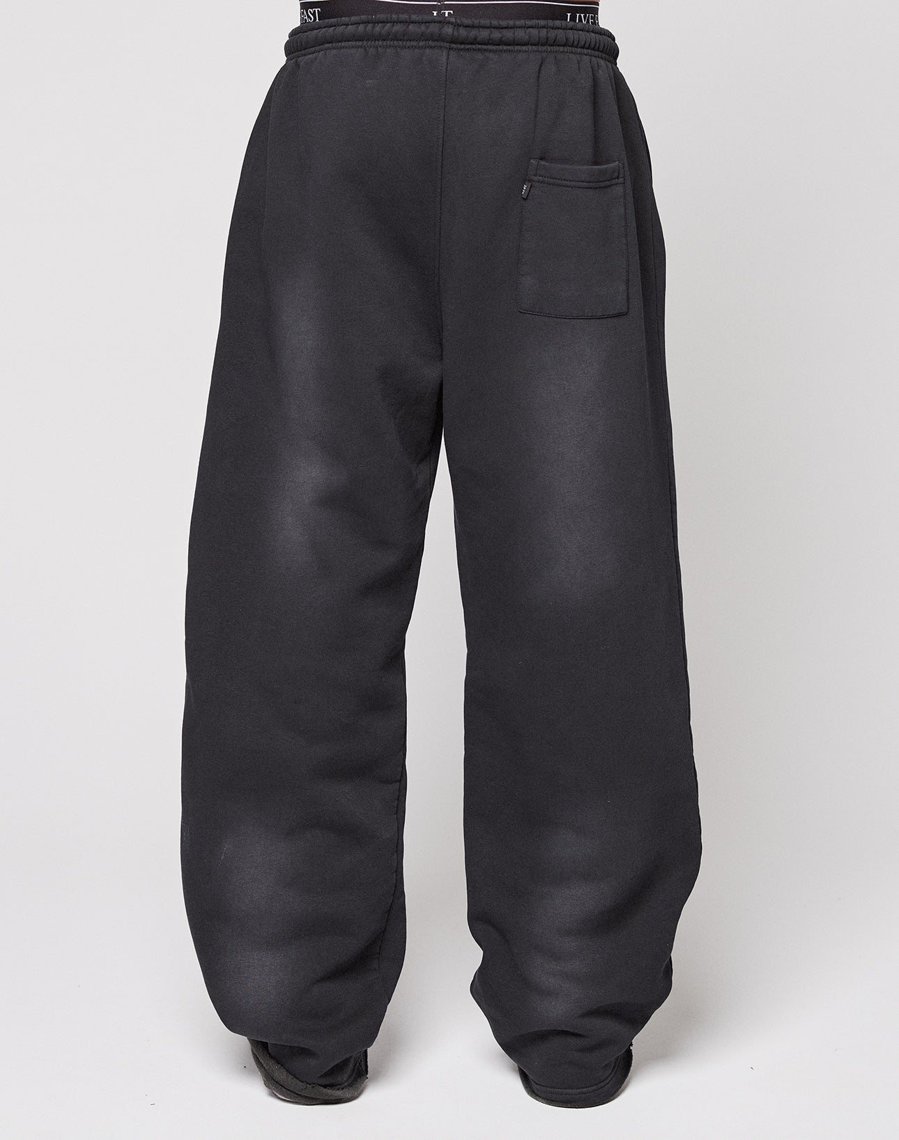 A person models the LIVE FAST DIE YOUNG Live Fast Rhinestone Jogger, featuring a wide fit, high waist, black color, and single front pocket. Their upper body is not visible as they stand against a plain white background.
