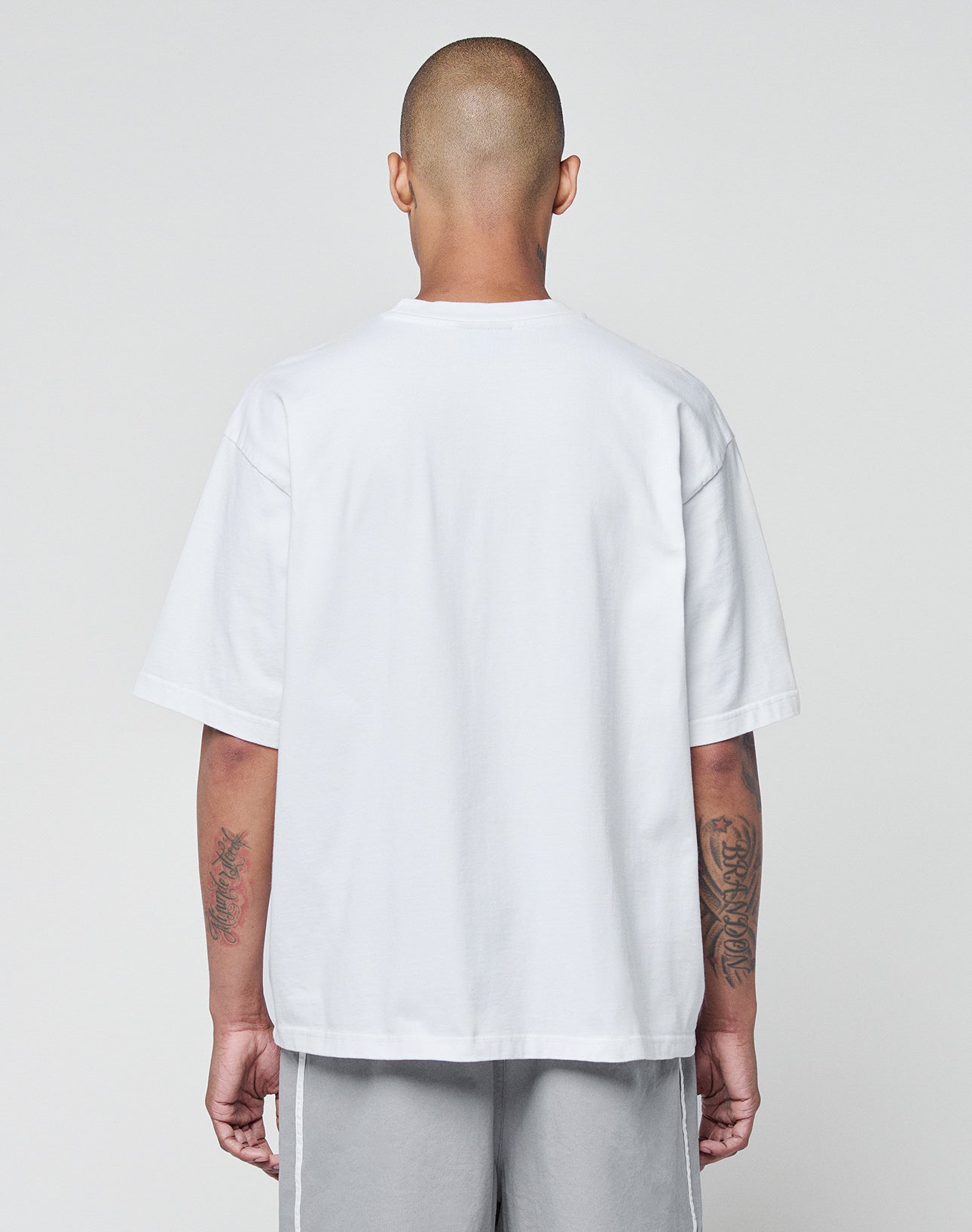 A person with a shaved head stands with their back to the camera, wearing a LIVE FAST DIE YOUNG Basic +49 Tee in oversize fit and light gray shorts. Both forearms are tattooed, set against a plain light gray background.