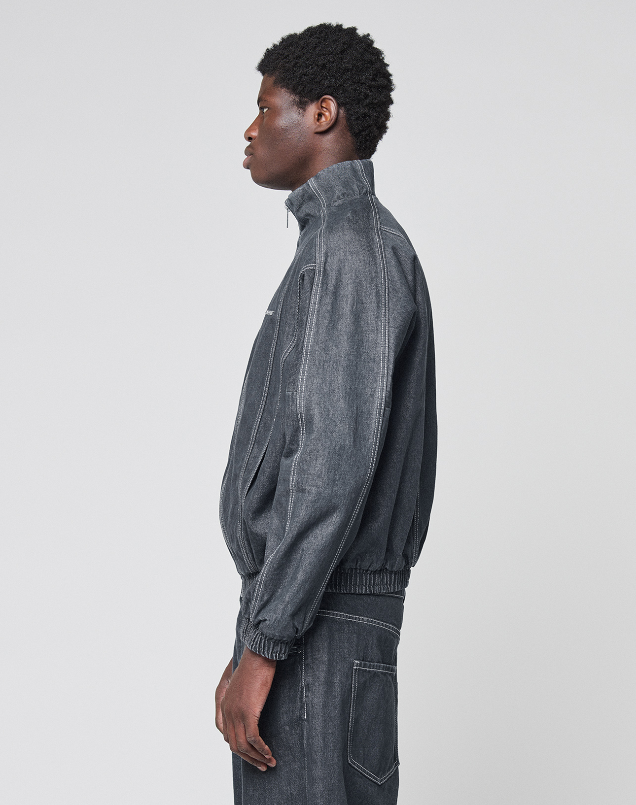 A person in profile wears the LIVE FAST DIE YOUNG LFDY Denim Trackjacket, a dark gray 100% Baumwolle jacket with Stehkragen, zip-up front, white stitching, elastic waistband and cuffs, paired with matching pants.
