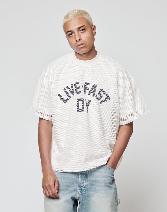 A person with short bleached blond hair wears the LFDY Boxy Jersey from LIVE FAST DIE YOUNG and light blue jeans, standing against a plain light gray background with a neutral expression.