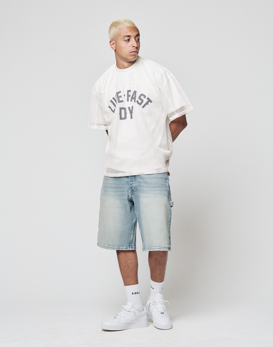 A man with short bleached blond hair wears the LFDY Boxy Jersey by LIVE FAST DIE YOUNG, light blue denim shorts, white socks, and white sneakers while standing against a plain white background.
