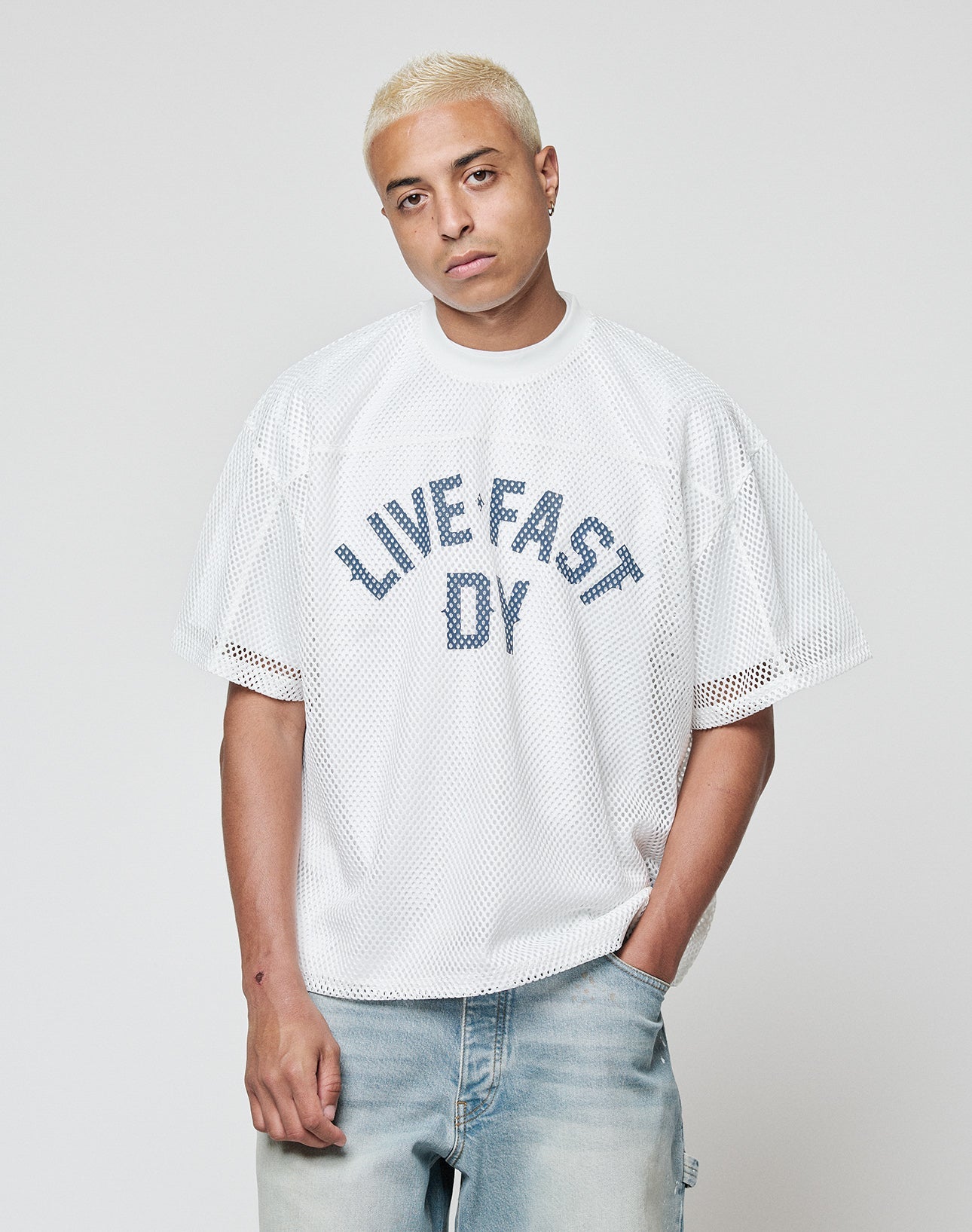 A person with short blond hair wears the LIVE FAST DIE YOUNG LFDY Boxy Jersey, a white mesh shirt with a bold front print, paired with light blue jeans, standing against a plain light background with one hand in their pocket.