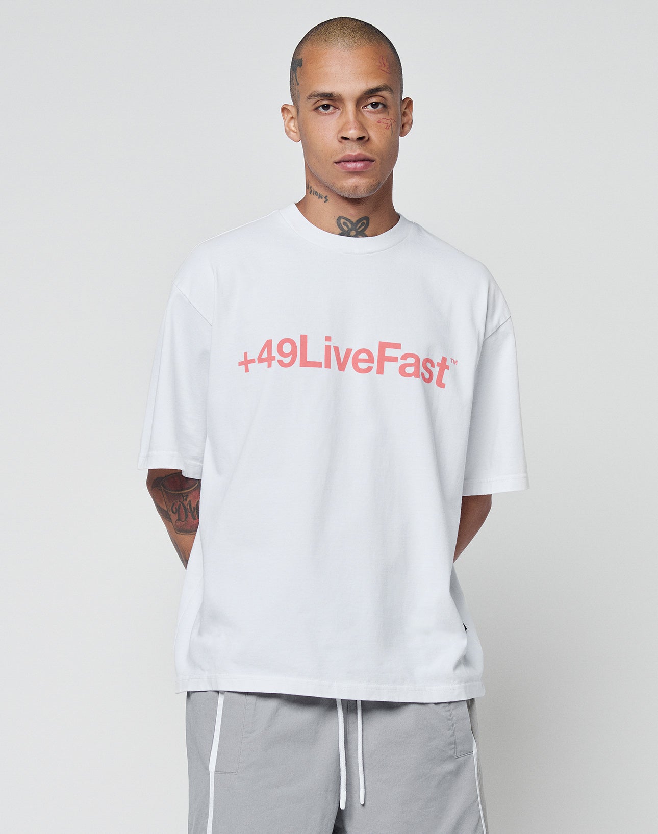A tattooed person stands against a plain backdrop, wearing the LIVE FAST DIE YOUNG Basic +49 Tee—an oversized white cotton shirt with red +49LiveFast print on the chest—and light gray shorts with white side stripes.