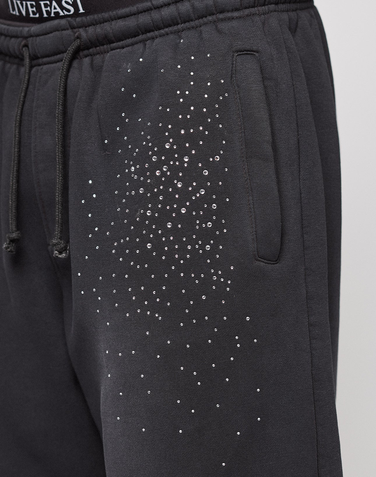 The Live Fast Rhinestone Jogger by LIVE FAST DIE YOUNG features black sweatpants with an elastic waistband, side pocket, rhinestone Muster on the upper thigh, partially visible "LIVE FAST" text, and a wide fit for comfort.