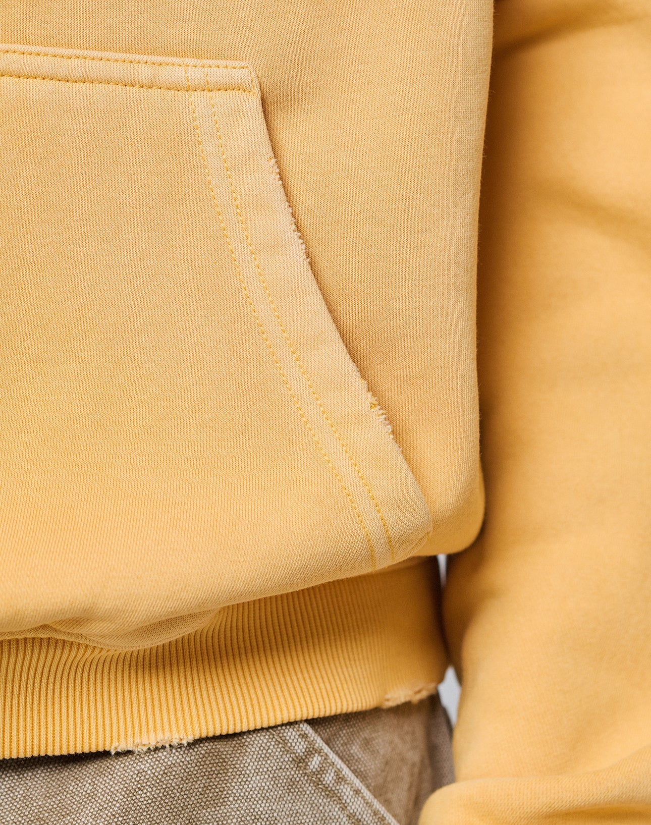 Close-up of a person wearing the LIVE FAST DIE YOUNG LFDY Corp. Hooded sweatshirt in light yellow, oversized fit with a front pocket and ribbed hem, paired with khaki pants. Only part of the arm, pocket, and waistband are visible.