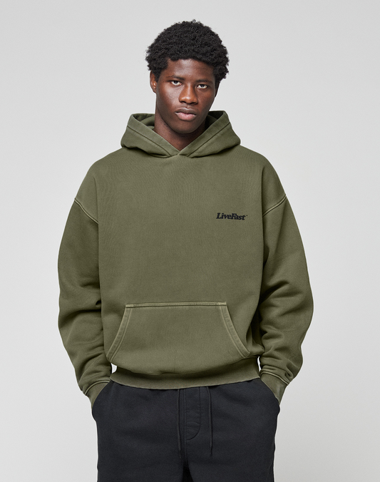 A young man with short curly hair models the LIVE FAST DIE YOUNG Basic 360 Hooded in olive green, featuring topstitching and an oversized fit, paired with black pants against a plain light gray background.