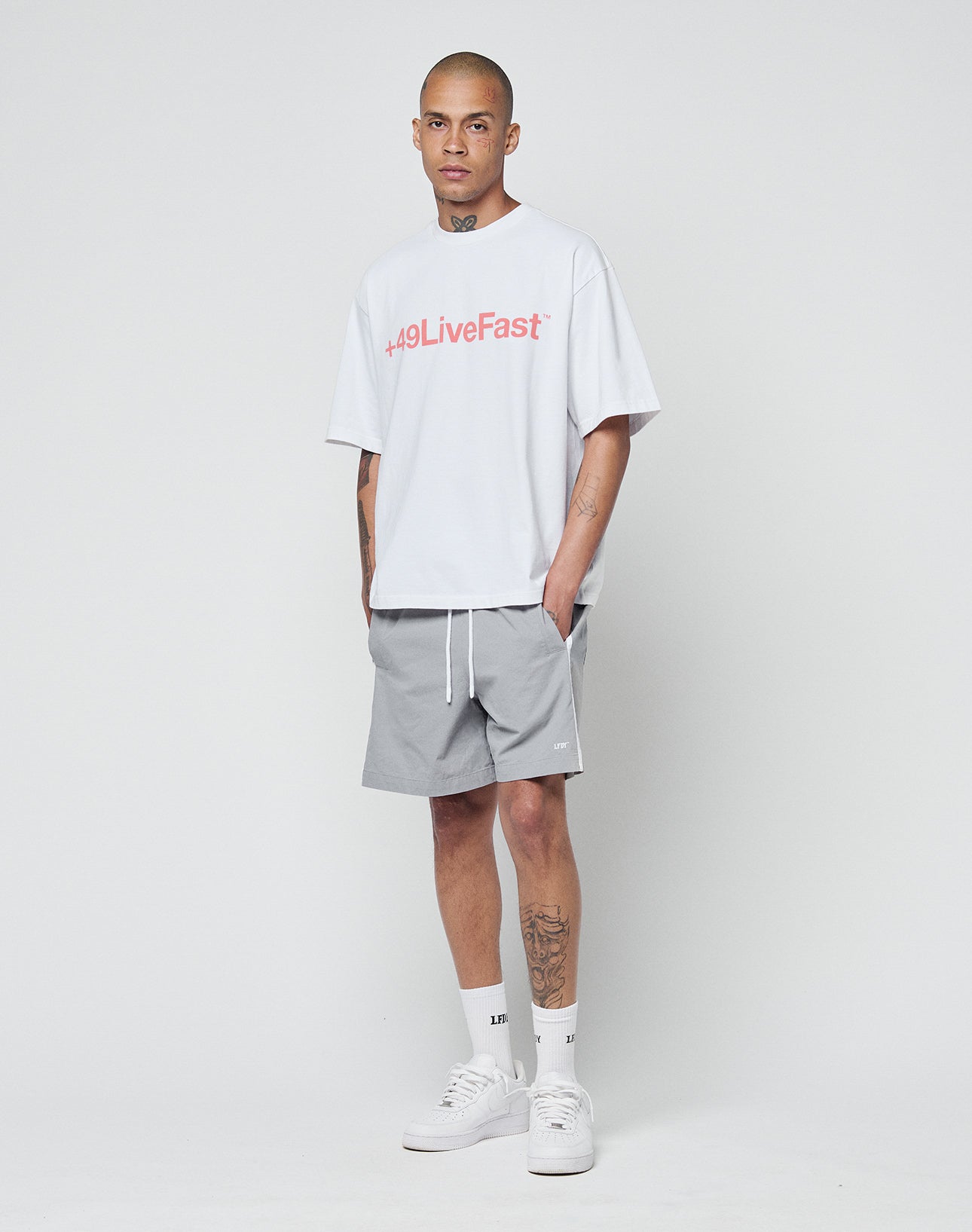 A person with tattoos stands against a plain white background wearing the LIVE FAST DIE YOUNG Basic +49 Tee in white with red frontprint, gray shorts, white crew socks, and white sneakers, showing a neutral expression.