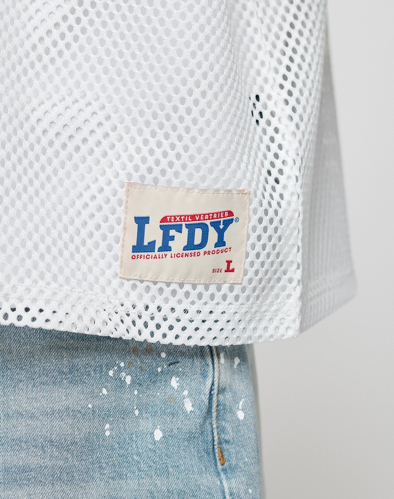 A close-up of the LFDY Boxy Jersey by LIVE FAST DIE YOUNG, a white mesh shirt with a rectangular LFDY OFFICIALLY LICENSED PRODUCT Size L tag, styled above blue jeans with white paint splatters.