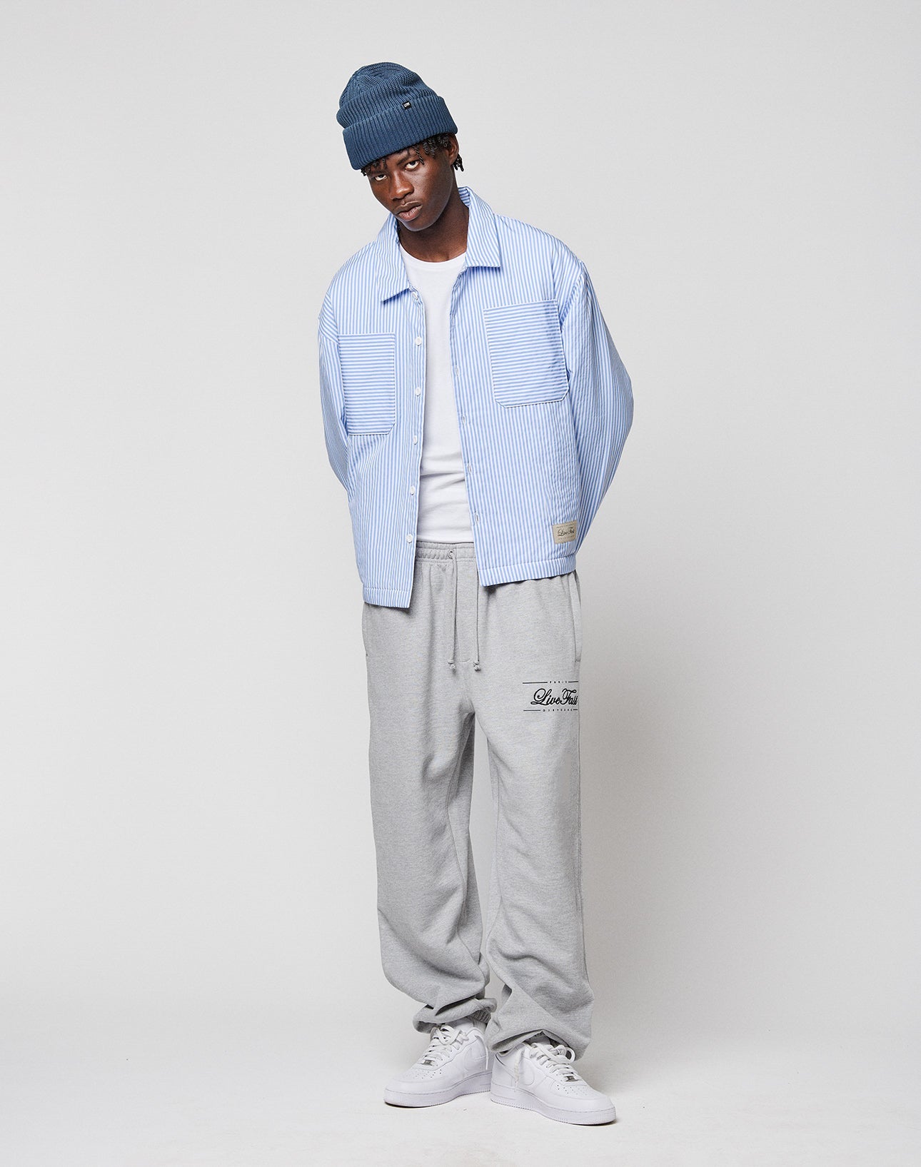 A man in a blue beanie, white T-shirt, gray sweatpants, white sneakers, and the LIVE FAST DIE YOUNG LFDY Striped Padded Shirt stands against a plain light background with his hands behind his back.