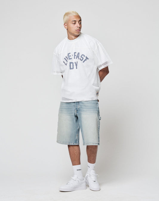 A man with short bleached blond hair wears the LFDY Boxy Jersey by LIVE FAST DIE YOUNG, featuring a bold frontprint, paired with light denim knee-length shorts, white socks, and white sneakers against a plain light background.