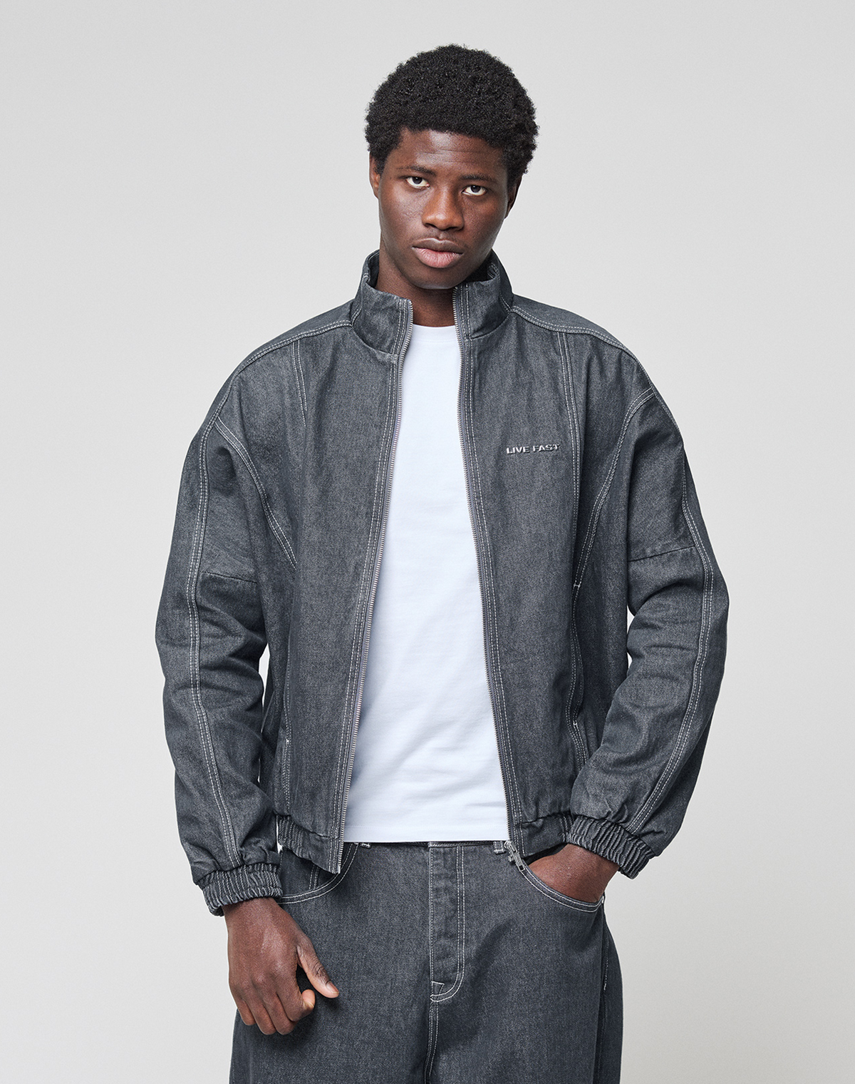 A person wears the LFDY Denim Trackjacket by LIVE FAST DIE YOUNG in dark gray over a white t-shirt and matching denim pants, standing against a plain light gray background and looking at the camera with a neutral expression.