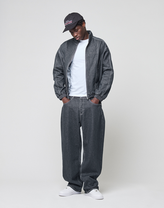 A person stands against a plain background, wearing the LFDY Denim Trackjacket by LIVE FAST DIE YOUNG with Stehkragen and Metall-Logo, matching baggy jeans (100% Baumwolle), a white T-shirt, white sneakers, and a black cap. Hands in pockets, head tilted down.