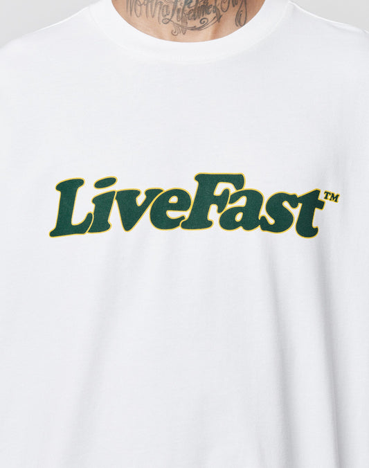A person wears the LIVE FAST DIE YOUNG Basic Tee, a white 100% cotton oversize shirt with LiveFast™ in bold dark green outlined in yellow on the front. A neck tattoo is partly visible above the shirt’s collar.