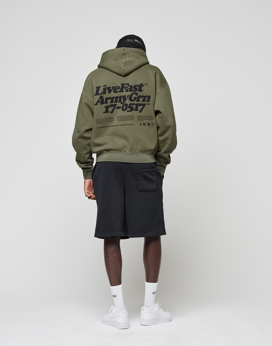 A person faces away, wearing the LIVE FAST DIE YOUNG Basic 360 Hooded in olive green with large black front and back print (oversize fit), black shorts, white socks, and white sneakers against a plain light gray background.