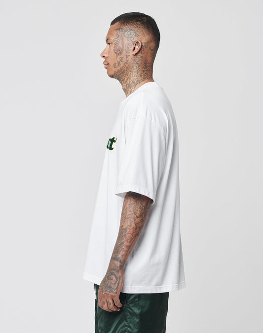 A tattooed man in profile wears the LIVE FAST DIE YOUNG Basic Tee, an oversize fit white t-shirt made from 100% Baumwolle, paired with dark green shorts against a plain light background.