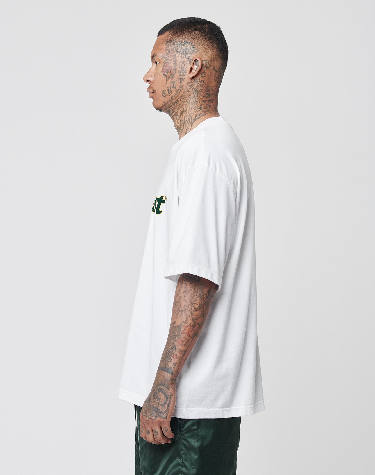 A tattooed man in profile wears the LIVE FAST DIE YOUNG Basic Tee, an oversize fit white t-shirt made from 100% Baumwolle, paired with dark green shorts against a plain light background.