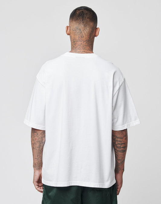 A tattooed man stands with his back to the camera, wearing the LIVE FAST DIE YOUNG Basic Tee in plain white and dark green shorts, against a light gray background.