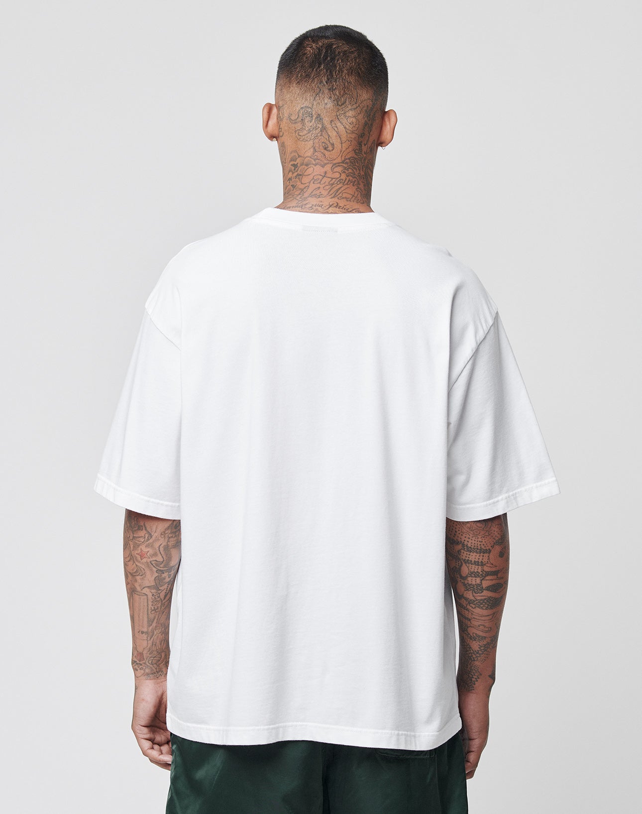 A tattooed man stands with his back to the camera, wearing the LIVE FAST DIE YOUNG Basic Tee in plain white and dark green shorts, against a light gray background.
