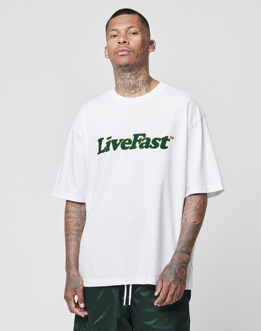 A person with tattoos on their neck and arms wears the LIVE FAST DIE YOUNG Basic Tee in white with LiveFast in green and yellow, paired with dark green shorts, standing against a plain light gray background.