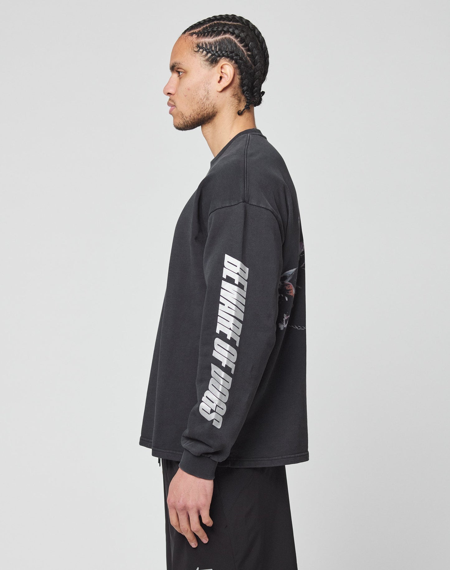 A man with cornrow braids stands in profile against a plain light gray background, wearing the LIVE FAST DIE YOUNG LFDY Canine Longsleeve in black and matching black pants, with reflective DETANGL OF DOGS print on the sleeve.