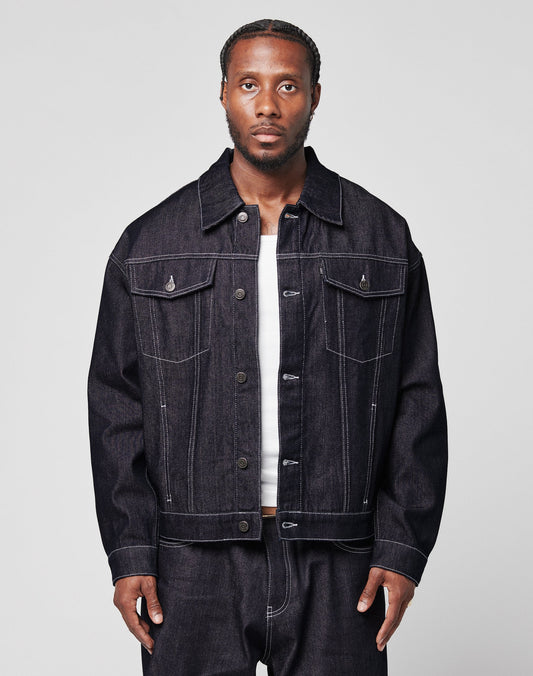 A man with braided hair wears an oversize LFDY Denim Jacket by LIVE FAST DIE YOUNG and matching raw-denim jeans over a white shirt, standing against a plain light gray background. | model