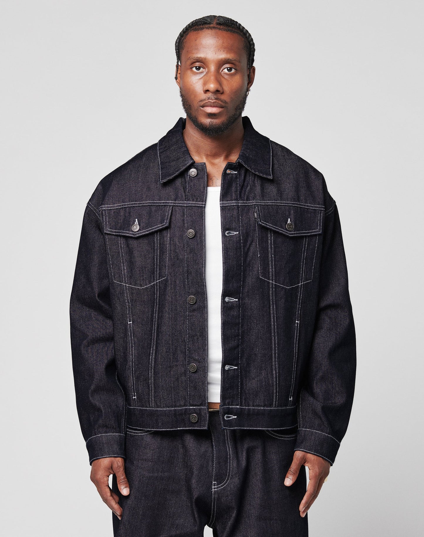 A man with braided hair wears an oversize LFDY Denim Jacket by LIVE FAST DIE YOUNG and matching raw-denim jeans over a white shirt, standing against a plain light gray background.