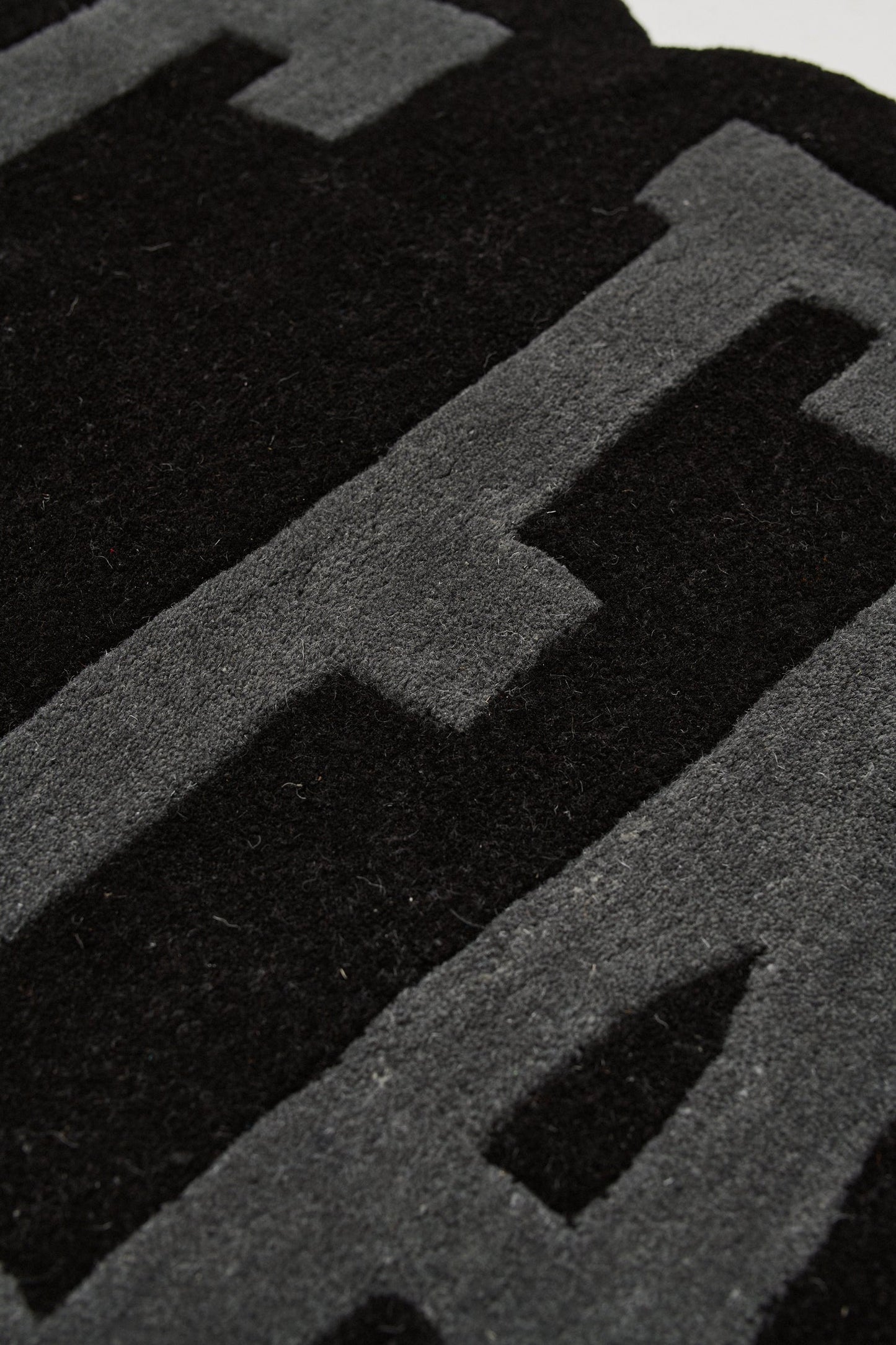 Close-up of the Live Fast Carpet by LIVE FAST DIE YOUNG, made from 100% Wolle in textured black and dark gray with bold block letters. Its soft surface suggests rug use; partial text visible. Designed for easy Teppichpflege.