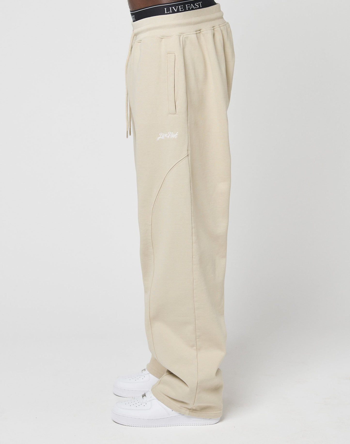 A side view of someone wearing beige Basic Wide Jogger pants from LIVE FAST DIE YOUNG with an elastic waistband and white sneakers. The LIVE FAST branded black underwear waistband is visible. The background is plain white.