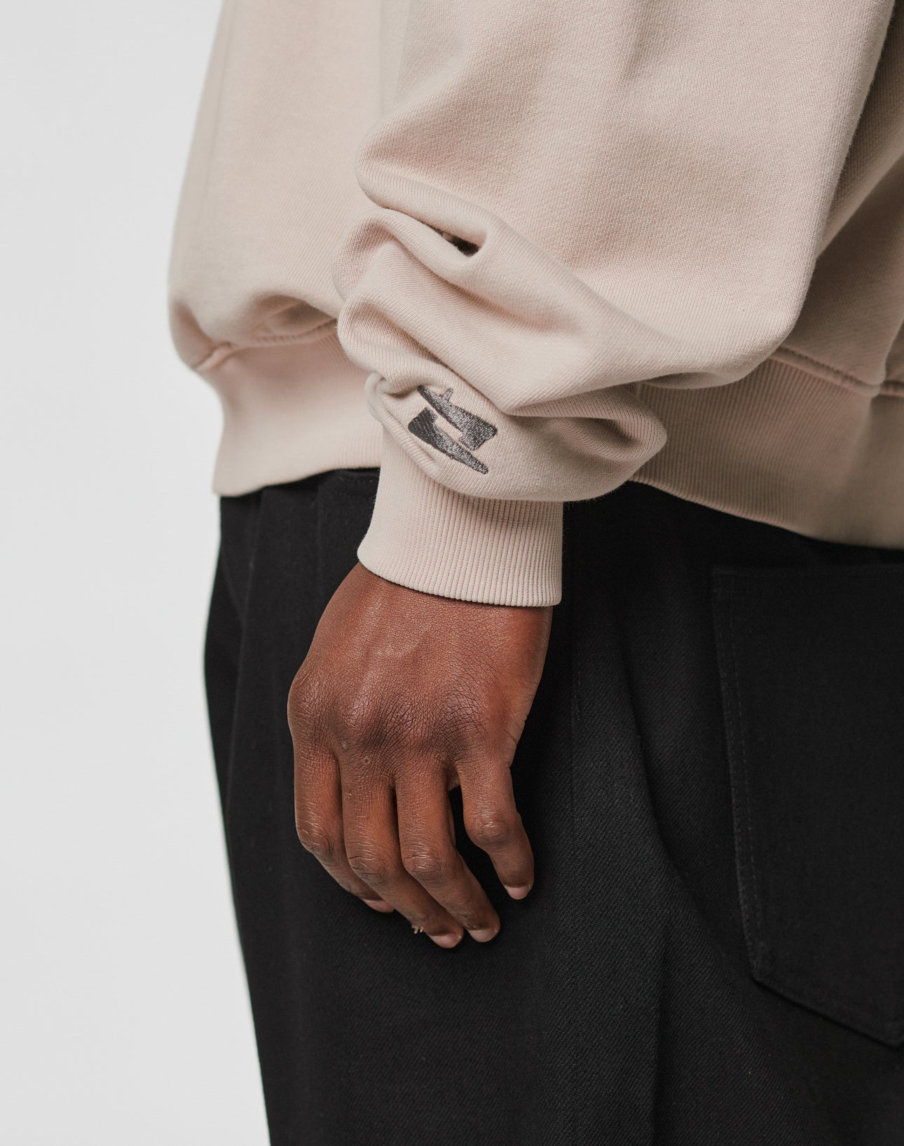 A close-up shows a hand resting by the side, wearing the LIVE FAST DIE YOUNG Live For It Sweater in beige with a small black graphic and Oversize Fit, paired with black pants with a large pocket against a plain light background.