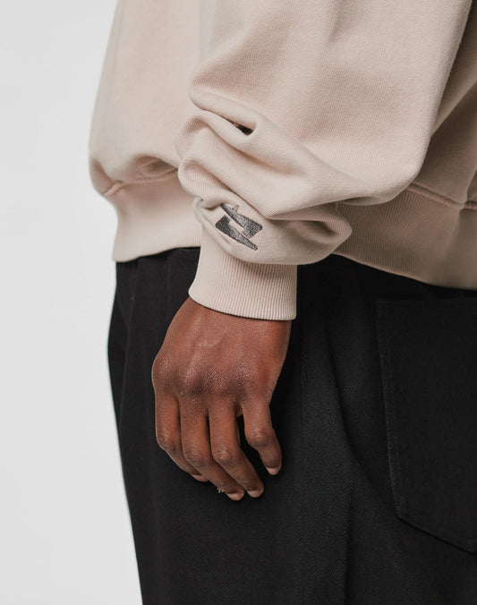 A close-up shows a hand resting by the side, wearing the LIVE FAST DIE YOUNG Live For It Sweater in beige with a small black graphic and Oversize Fit, paired with black pants with a large pocket against a plain light background.