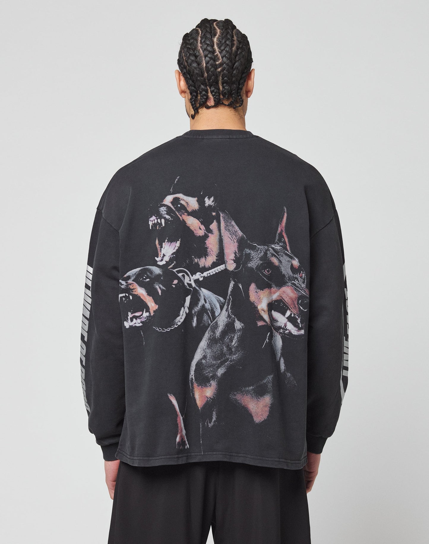 A person with braided hair faces away, wearing the LIVE FAST DIE YOUNG LFDY Canine Longsleeve—an oversize-fit black shirt made from 100% cotton, featuring three barking Dobermans on the back and light stripes on the sleeves.