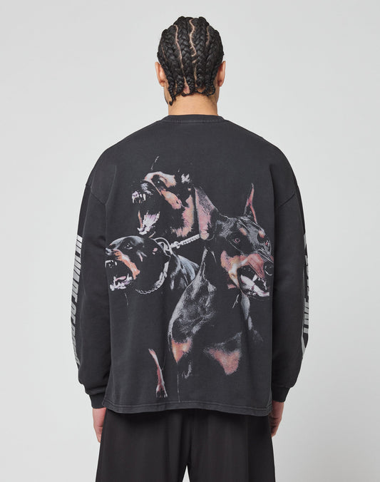A person with braided hair faces away, wearing the LIVE FAST DIE YOUNG LFDY Canine Longsleeve—an oversize-fit black shirt made from 100% cotton, featuring three barking Dobermans on the back and light stripes on the sleeves.