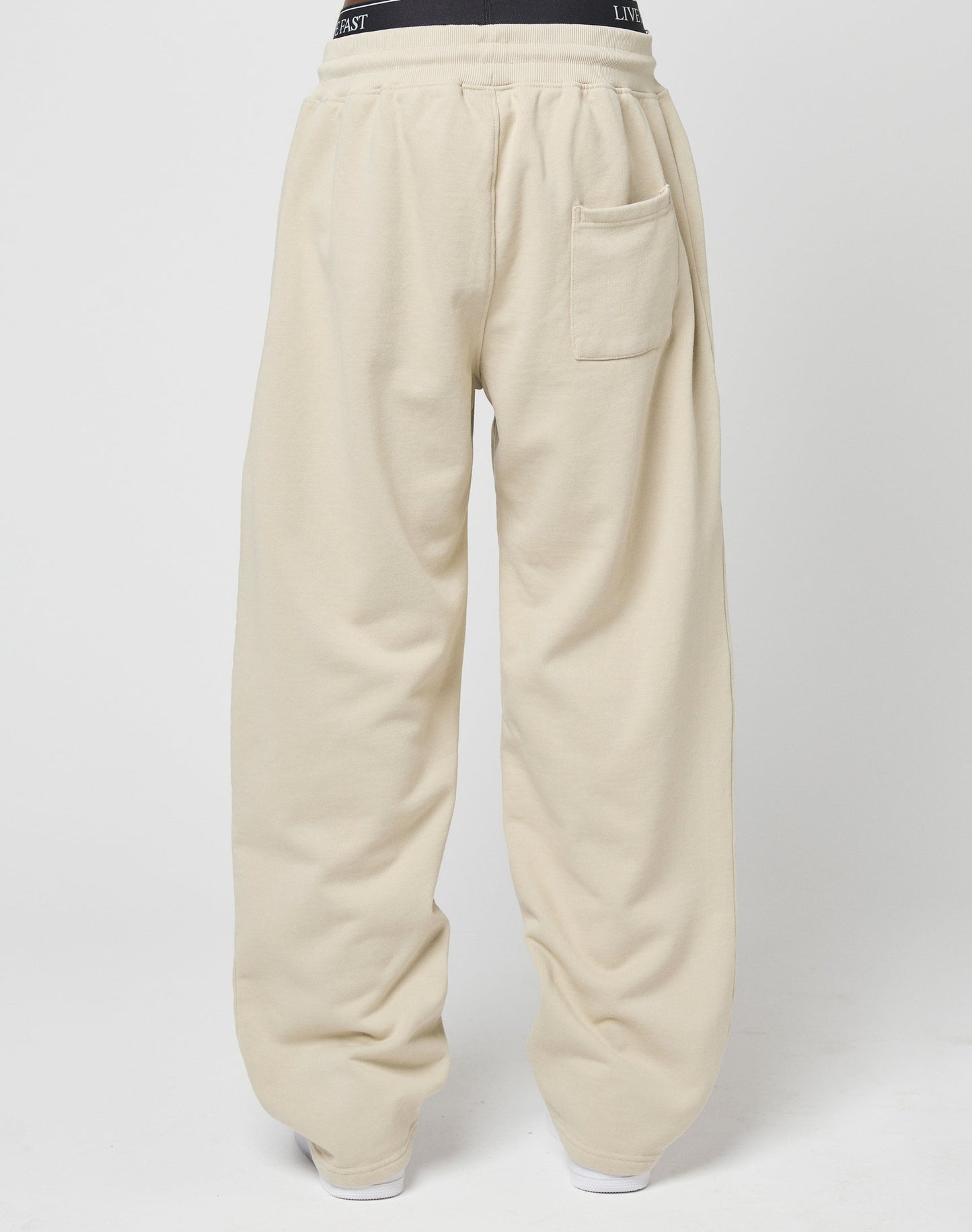A person wearing LIVE FAST DIE YOUNG Basic Wide Jogger in beige wide fit, featuring a single back pocket and partially visible black Elastikbund waistband, shown from behind against a plain white background.
