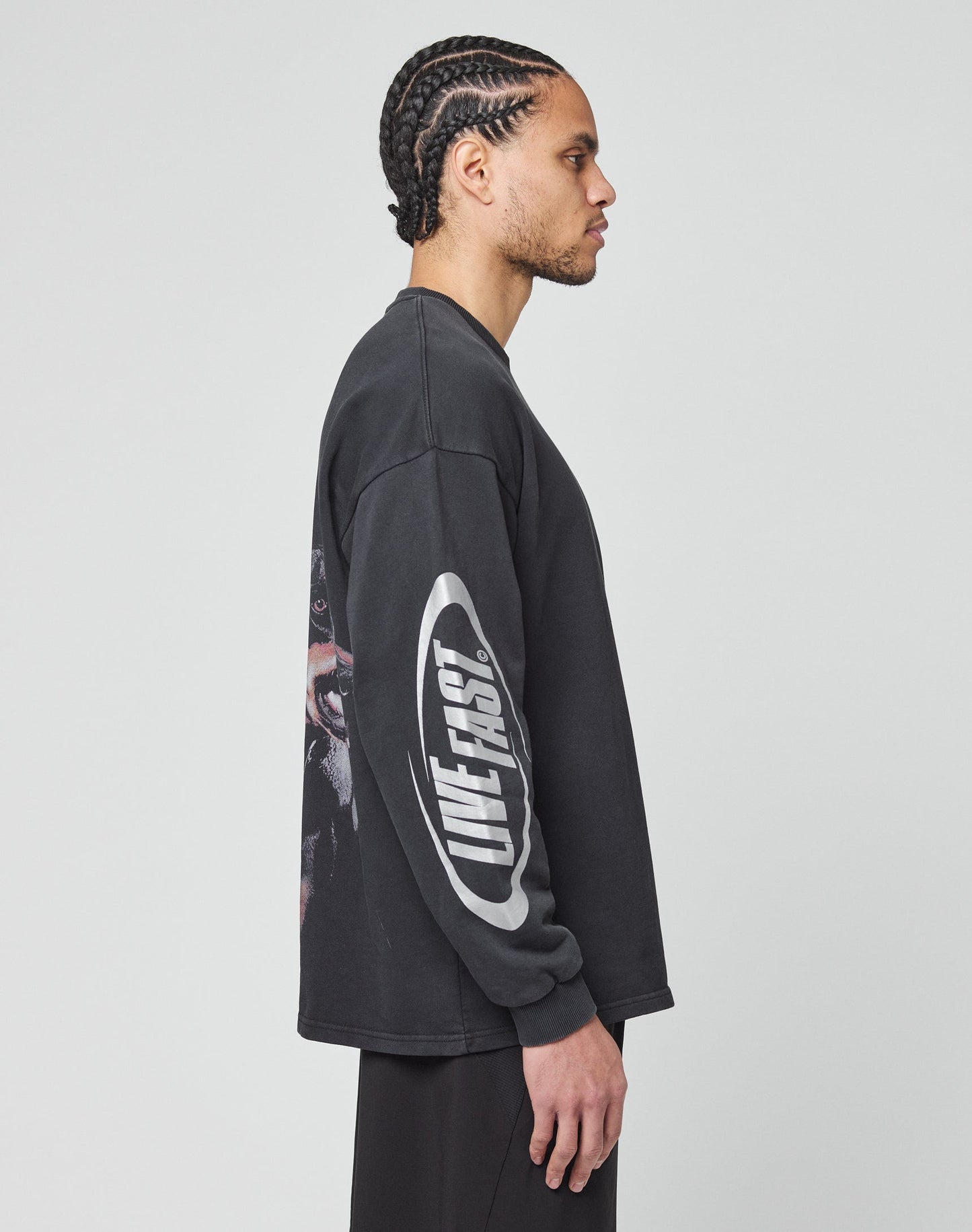 A man with braided hair stands in profile wearing an oversize black LFDY Canine Longsleeve by LIVE FAST DIE YOUNG, featuring bold white LIVE FAST lettering inside an oval on the sleeve. The background is plain and light gray.