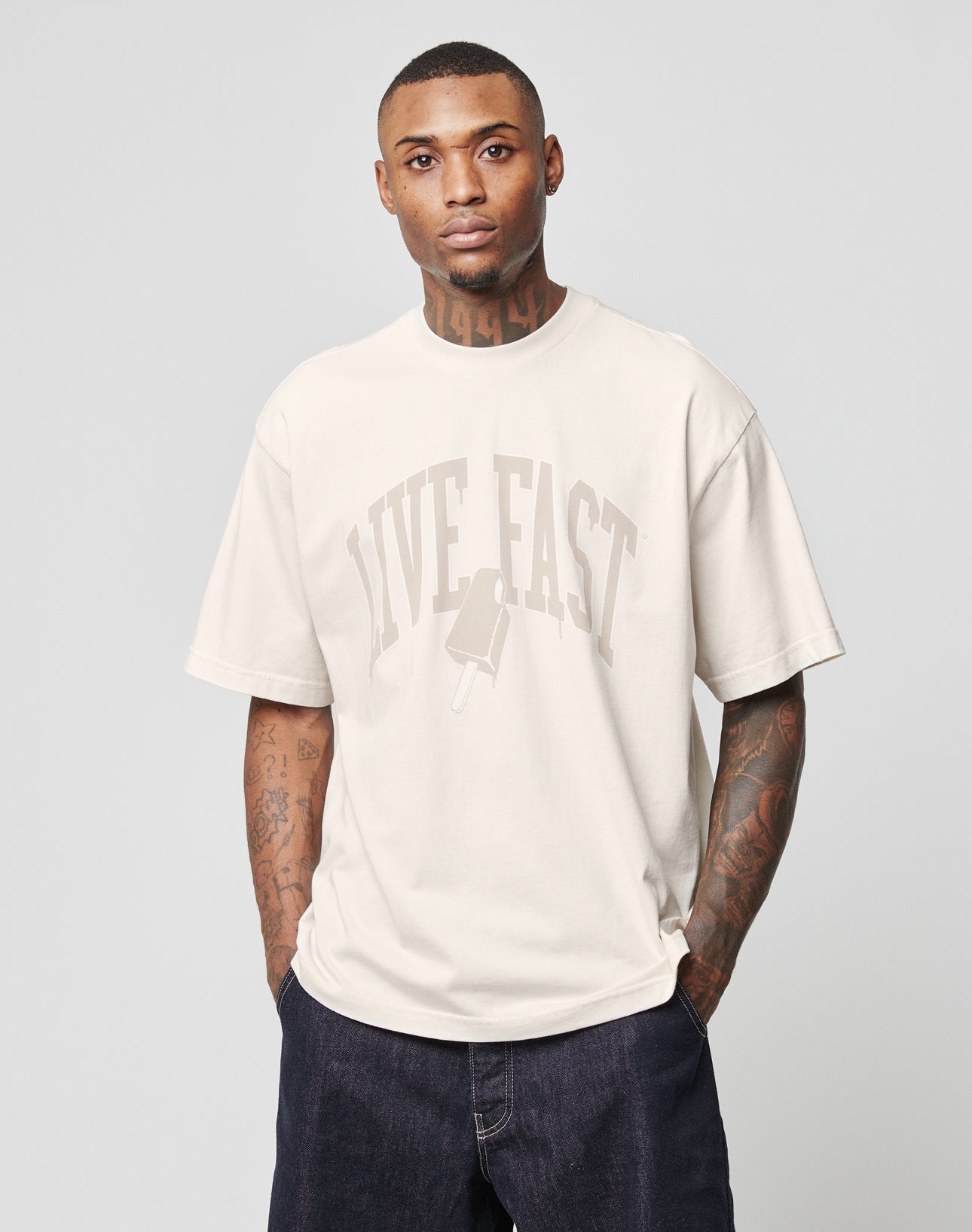 A man poses against a plain background in the LIVE FAST DIE YOUNG Live Fast Ice Tee, featuring “LIVE FAST” and a popsicle graphic on the front, styled with dark denim jeans.
