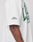 A person wears the LIVE FAST DIE YOUNG Live Fast Stallion Tee—an oversized white shirt with green front print and emblems on chest and sleeve, revealing a detailed arm tattoo. The background is light and plain.